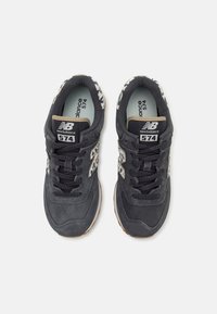 Ζευγάρι μαύρα αθλητικά παπούτσια New Balance 574 με λευκά σχέδια λεοπάρδαλης και μαύρα κορδόνια, από ψηλά, σε λευκό φόντο.