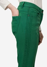 Zielone dżinsy z boczną kieszenią i widocznymi detalami szwów. Tkanina jest gładka z lekko chropowatym wykończeniem.