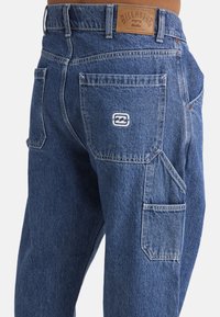 Denim cargobroek in medium blauw met een gestructureerde afwerking, voorzien van een leren logopatch en meerdere zakken voor functionaliteit.