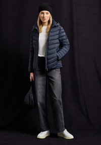 Veste rembourrée navy avec fermeture éclair et capuche, associée à un pull léger, un jean foncé et des baskets blanches ; tenant un petit sac noir.