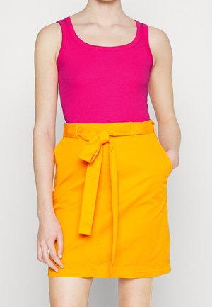 Femme portant un haut sans manches rose vif et une jupe jaune taille haute avec une ceinture nouée et des poches latérales.