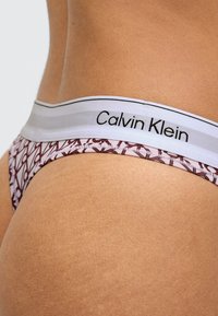 Detailný záber na osobu nosiacu vzorované spodné prádlo Calvin Klein s bielym pásikom v páse, na ktorom je zobrazený názov značky.