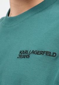 Tyrkysová mikina s okrúhlym výstrihom a rebrovaným golierom, s čiernym vyšitým textom "KARL LAGERFELD JEANS" na prednej strane.