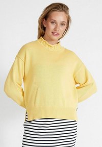 Gelber gestrickter Pullover mit einem gerafften Ausschnitt und langen, lockeren Ärmeln. Getragen über einem schwarz-weißen gestreiften Rock.
