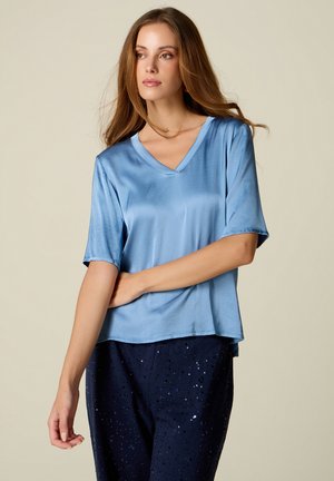 Mujer con cabello largo y castaño lleva un top de satén azul claro con escote en V y pantalones azul oscuro con lentejuelas, posando frente a un fondo beige.