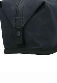 Esquina de una bolsa de tela negra con un acercamiento a un botón a presión negro de marca y un detalle de correa tejida reforzada.