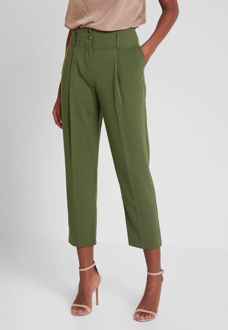 Pantalones verdes de cintura alta con pliegues frontales, dos botones y bolsillos laterales. Largo recortado con una textura de tela lisa y estructurada.