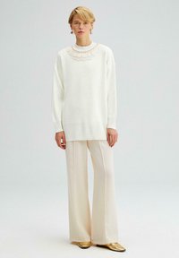 Witte gebreide trui met een kanten kraag met golven, gecombineerd met lichtbeige wijde broek en gouden schoenen, tegen een neutrale achtergrond.