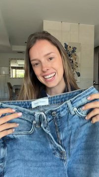 Femeie zâmbitoare ținând o pereche de blugi albaștri cu fermoar deschis într-un cadru modern de interior.