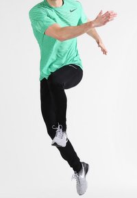 Camisa deportiva Nike de color verde claro con un patrón texturizado, combinada con leggings negros y zapatillas de correr grises, mostrando una acción de salto.