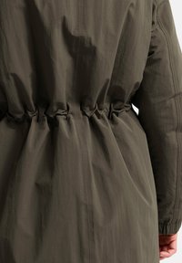 Manteau léger vert olive avec taille cintrée. Texture lisse, manches longues et fronces élastiques à la taille pour une silhouette ajustée.