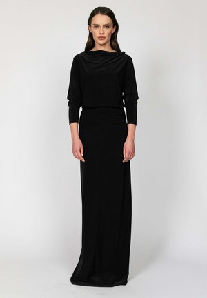 Kobieta o długich brązowych włosach stoi przodem, ubrana w długą czarną sukienkę z długimi rękawami na jasnym, jednolitym tle.