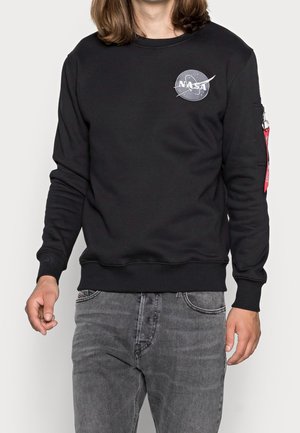 Mężczyzna w czarnej bluzie z logo NASA z czerwoną metką na kieszeni lewej rękawicy i wyblakłych szarych dżinsach, stojący na białym tle.