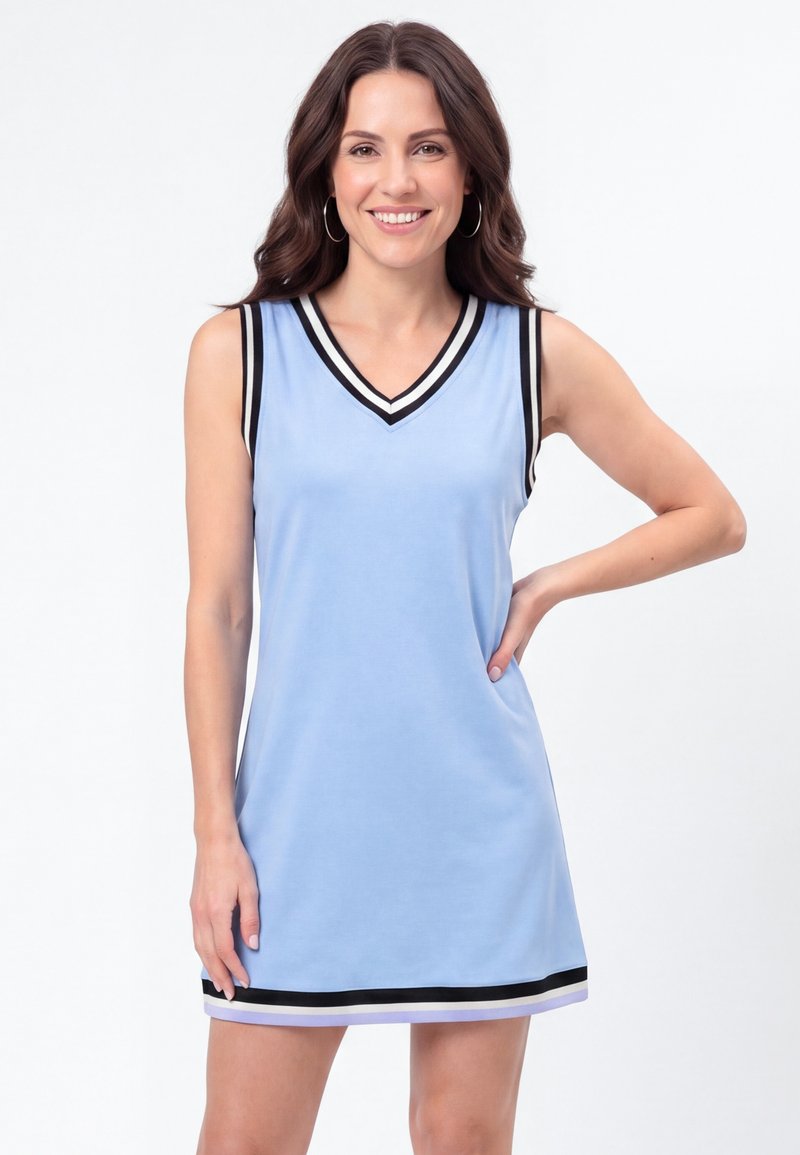 Femme aux cheveux bruns portant une robe bleu clair sans manches avec des bordures noires et blanches, souriante, une main sur la hanche.