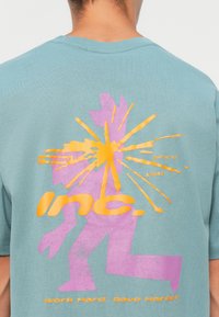 Light blue cotton t-shirt featuring a large, abstract graphic in pink and orange on the back. Text reads "inc. since 1981" and "Work Hard. Rave Harder."