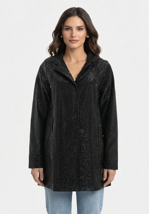 Femme portant une veste noire à capuche avec motif et une fermeture éclair devant, sur un jean bleu clair, debout sur un fond blanc uni.