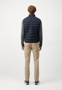 Marineblauer Steppwesten mit Steppmuster, hoher Kragen, über einem grünen Langarmhemd getragen. Kombiniert mit beigen Hosen und schwarzen Sneakern.