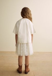 T-shirt en coton blanc superposé sur une robe en dentelle blanche, associé à des bottes à la cheville brunes. La tenue a une coupe décontractée et des accents texturés.