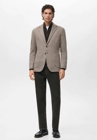 Blazer beige con rever a V, due tasche frontali e pantaloni grigio scuro. Il modello indossa un maglione scuro sotto, abbinato a scarpe nere.