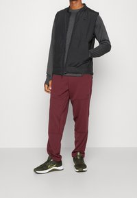 Schwarze Sportweste über einem grauen Langarmshirt, kombiniert mit kastanienbraunen Hosen mit strukturiertem Muster und grünen Sneakers mit gelben Akzenten.