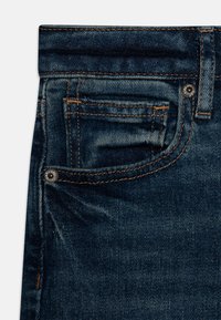 Jeans de mezclilla azul oscuro con diseño clásico de cinco bolsillos, costuras visibles y un pequeño remache de metal en el bolsillo delantero.