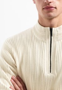 Cream ribbed knit pullover with a high collar and black zipper. Notable texture and pattern create a structured appearance.