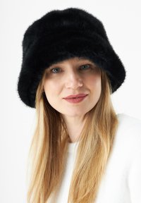 Zwarte faux fur bucket hat met een ronde bovenkant, zachte textuur en brede rand. Model draagt een witte sweater, medium lang blond haar zichtbaar.