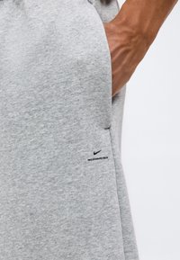 Gros plan d'une main dans la poche d'un pantalon de survêtement Nike gris clair avec le logo "Nike Standard Issue" en noir près de la poche.