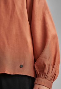 Rostfarbener Langarmbluse mit gerafften Bündchen und schwarzen Hosen, die unten sichtbar sind, vor einem einfarbigen grauen Hintergrund.