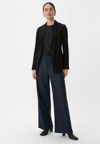 Blazer noir ajusté sur un blouse foncée et volantée, associé à un jean en denim foncé à jambes larges et des bottines à talons noires.
