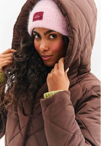 Žena s kučeravými vlasmi, ktorá má na hlave ružovú pletenú čiapku a hnedú prešívanú bundu, drží kapucňu bundy blízko tváre.