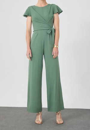 Kobieta ubrana w oliwkowo-zielony kombinezon z rozkloszowanymi rękawami, wiązany w talii, szerokimi nogawkami, złotą bransoletką i złotymi sandałkami na obcasie.