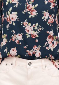 Blouse bleu marine à motif floral rose et rouge, rentrée dans un jean taille haute rose clair avec bouton métallique et rivets.