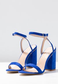 Pari av亮藍色絨面高跟涼鞋，配有腳踝帶和方塊跟，放置在白色表面和灰色背景上。