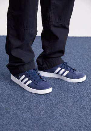 Personne portant des baskets bleu marine avec des bandes blanches et des semelles blanches, debout sur un sol recouvert d'un tapis bleu.