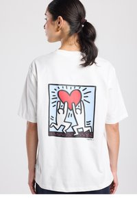 Wit katoenen T-shirt met een grote afbeelding op de achterkant van twee gestileerde figuren die een rood hart vasthouden tegen een lichtblauwe achtergrond.