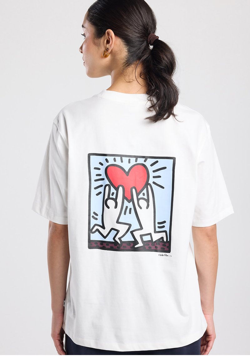 Wit katoenen T-shirt met een grote afbeelding op de achterkant van twee gestileerde figuren die een rood hart vasthouden tegen een lichtblauwe achtergrond.