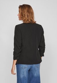 Femme aux cheveux mi-longs portant un blazer noir à manches fronçées et un jean bleu, vue de dos sur un fond uni.