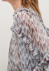 Donna che indossa una blusa trasparente con motivo, con dettagli arricciati lungo la spalla e la manica, mostrata di lato.