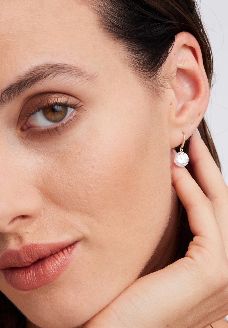 Złote kolczyki w kształcie dużego okrągłego kryształu, prezentujące błyszczącą powierzchnię i elegancki design. Noszone na uchu modelki.