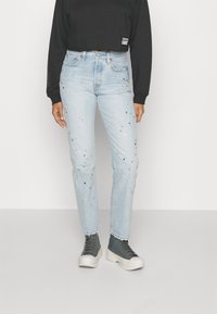 Vaqueros de mezclilla azul claro con detalles desgastados y adornos plateados y azules, combinados con una blusa negra de manga larga y zapatillas grises.