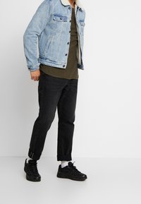 Giacca di jeans blu chiaro con colletto in shearling, t-shirt verde oliva, pantaloni neri a gamba stretta e sneakers nere con calzini bianchi.