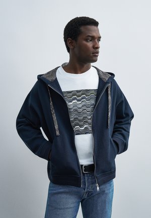 Mężczyzna noszący granatową bluzę z kapturem na zamek błyskawiczny na białej koszulce z czarnym, szarym i zielonym wzorem w zygzak, w połączeniu z niebieskimi dżinsami i rękami w kieszeniach.