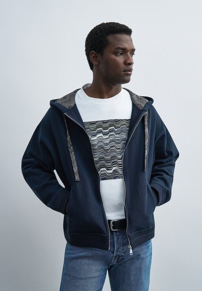 Homme portant un sweat à capuche zippé bleu marine sur un t-shirt blanc avec un motif en zigzag noir, gris et vert, associé à un jean bleu et les mains dans les poches.