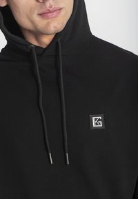 Sudadera negra de tela suave con capucha ajustable, que presenta un pequeño logo cuadrado en el pecho y terminales plateados en los cordones.