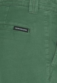 Gros plan sur un jean vert montrant un passant de ceinture, une poche avant et une étiquette noire Calvin Klein Jeans cousue au-dessus de la poche.