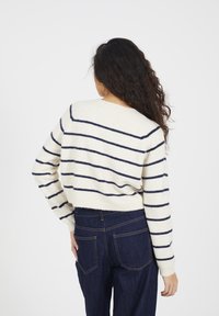 Donna con lunghi capelli ricci che indossa un maglione crema con strisce orizzontali blu navy e jeans blu scuro, in piedi contro uno sfondo bianco.