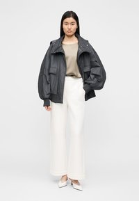 Juodos spalvos, per didelis švarkas su aukšta apykakle ir didelėmis kišenėmis, derinamas su smėlio spalvos viršutine dalimi ir baltomis plačiomis kelnėmis. Lygus tekstūros audinys ir laisvas siluetas.