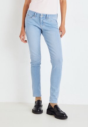 Person iført lyseblå slim-fit jeans, beige sokker og sorte læderloafers, stående mod en ensartet hvid baggrund.