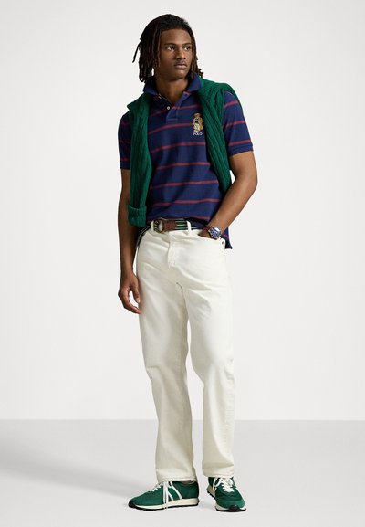 Marineblå og røde stribede polo-shirt med logo, hvide jeans, grøn strikket vest draperet over skuldrene og grønne sneakers.